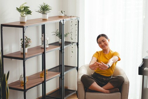 Asian woman sitting on armchair in living room relaxing stretching body breathing fresh air with trees feeling good stress relief at home.