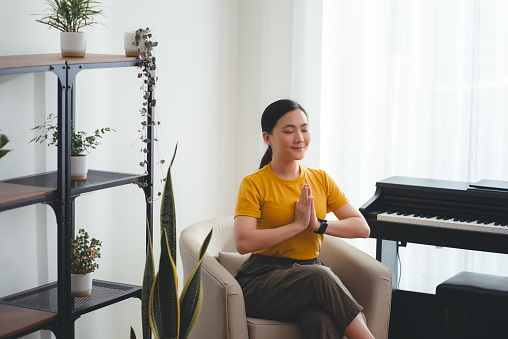 Asian woman with yoga practice closed eyes practicing breathing feel comfort do meditation practice in living room at home.