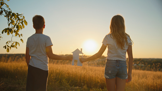 As The Sun Sets,Boy And Girl Gently Hold A Paper House Template In Their Hands Amidst A Peaceful Meadow