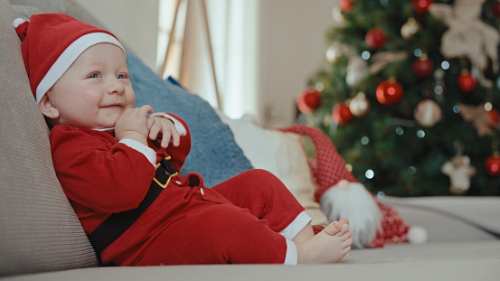 With a Contagious Smile,the Cute Baby Boy in a Santa Claus Costume Sits Comfortably on the Sofa at Home,Spreading Joy and Warmth Throughout the Festive Atmosphere.