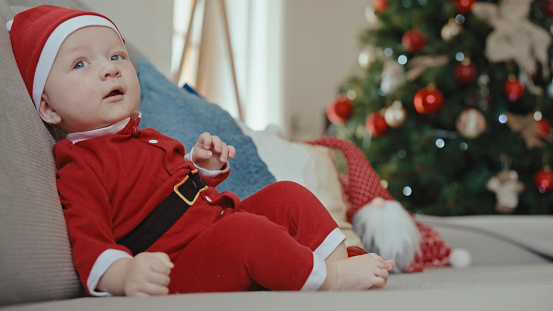 The Adorable Baby Boy,Dressed in a Santa Claus Costume,Sits on the Sofa at Home,Looking Away with Curiosity,His Innocence Captivating the Room with Charm and Sweetness.