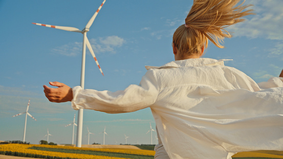 Woman Extending her Arms as She Joyfully Running through an Agricultural Farm,Heading Eagerly towards Colossal Wind Turbine Standing amidst the Countryside.