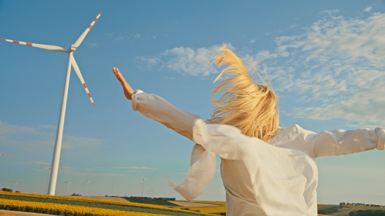 Amidst the Serene Expanse of an Agricultural Landscape,Woman with Outstretched Arms Racing towards Towering Wind Turbine with Boundless Energy and Enthusiasm in the Rural Setting.