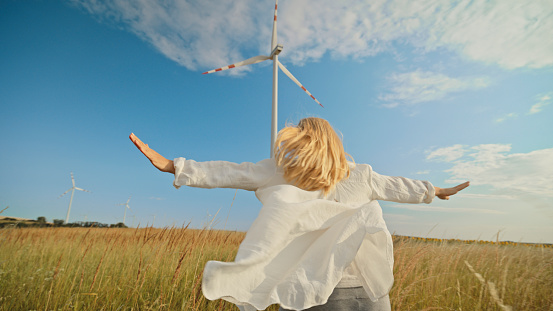 Woman in Casual Attire Joyfully Running to Colossal Wind Turbines in the Vast Agricultural Field,under the Bright Blue Sky on a Sunny Day.