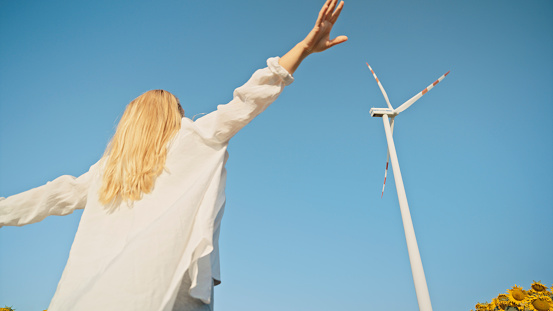 Woman Standing in Front of Towering Wind Turbine,Embracing the Gentle Caress of the Wind Flowing through her Fingers,Connecting her with the Energy of the Environment.