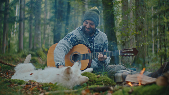 Holding his Guitar with a Contented Smile,a Happy Man Enjoys Playful Moments with his Dog as they Wait by the Campfire where Food is being Prepared,Savoring the Warmth of Companionship and the Rustic Charm of the Forest.