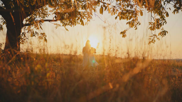 In the Golden Light of Sunset,a Mother Carries Her Baby while Sitting Under a Tree in the Field,the Peaceful Surroundings Fostering Moments of Connection and Warmth.