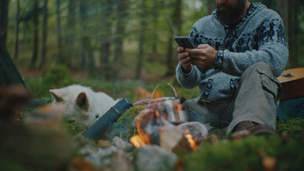 istock Man Using Mobile Phone while Food being Prepared on Campfire in Forest during Vacation 2025398424