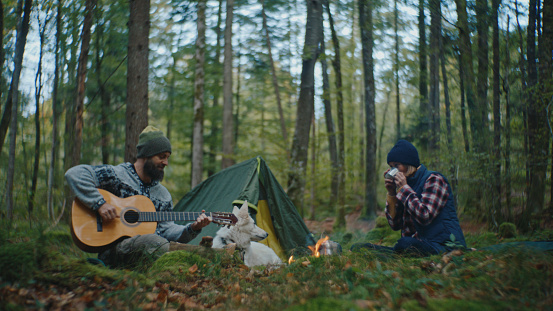 A Man Plays Guitar while a Woman Enjoys a Drink amidst the Forest during Vacation,their Leisurely Activities Enhancing the Natural Beauty and Serenity of the Woodland Surroundings.