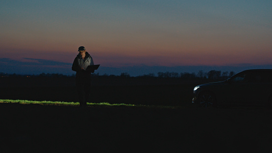 In The Peaceful Twilight Of The Agricultural Field's Blue Hour,An Agronomist Sits By His Car,Absorbed In His Laptop's Glow. He Seamlessly Blends Technology With Nature,Navigating Data To Optimize Farming Practices