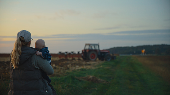 As The Sun Sets,A Female Farmer Holds Her Baby Boy Close,Observing A Tractor Plowing The Fields Against The Backdrop Of The Evening Sky. Heartwarming Scene Portrays A Mother's Nurturing Presence Amidst The Agricultural Work