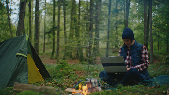 Amidst the Tranquil Woodland,a Woman Diligently Tends to her Laptop Tasks while the Campfire near the Tent Crackles with Warmth and the Scent of Cooking Food,Blending the Comforts of Technology with the Rustic Charm of Outdoor Living.