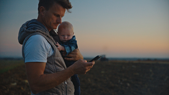 Farmer Father Stands On The Field,Holding His Baby Boy,And Managing His Responsibilities With A Mobile Phone. Showcasing The Farmer's Dedication To Both Nurturing The Next Generation And Tending To The Land