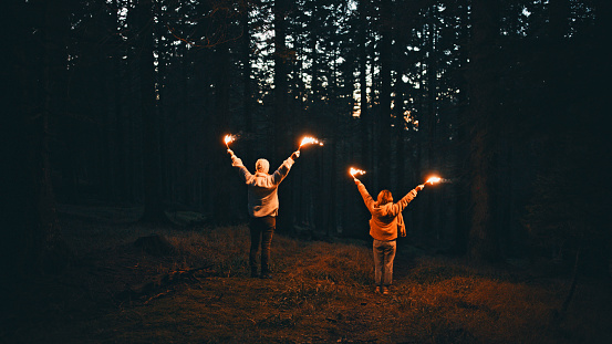 In the Darkness of the Forest at Night,a Rear View Captures Female Friends Holding Sparklers,Their Glowing Light Illuminating the Surroundings with a Warm and Magical Ambiance. Against the Backdrop of the Night Sky,Their Silhouettes and the Flickering Lights Create a Scene of Joy,Camaraderie and Enchantment