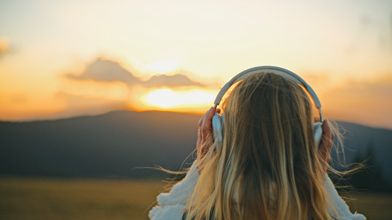 In the Enchanting Glow of Sunset,a Rear View Reveals a Young Woman with Blond Hair,Immersed in Music as She Listens Through Headphones against the Vibrant Sky. Her Silhouette against the Twilight Backdrop Exudes a Sense of Tranquility and Serenity