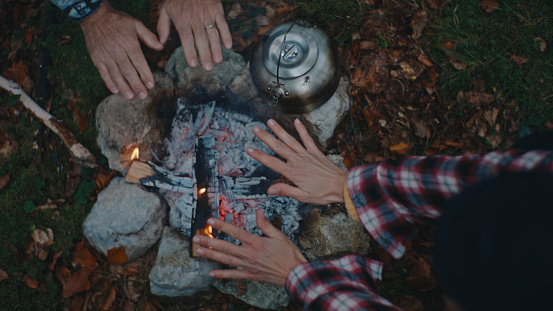 With a High Angle Perspective,a Couple Warms their Hands From the Burning Wood at the Campfire in the Forest,Finding Solace and Comfort in the Flickering Flames amidst Nature's Embrace.