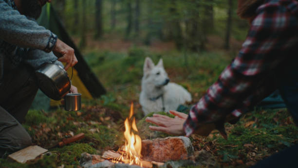 istock Man Pouring Drink in Cup while Woman Warming Hands in Campfire at Forest 2025402730