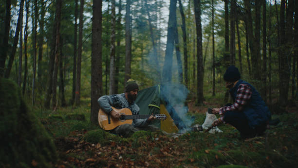 istock Man Playing Guitar while Woman Cooking Food on Campfire in Forest during Vacation 2025403664