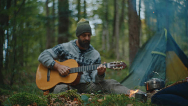 istock Man Playing Guitar while Woman Food being Prepared on Campfire in Forest during Vacation 2025403830
