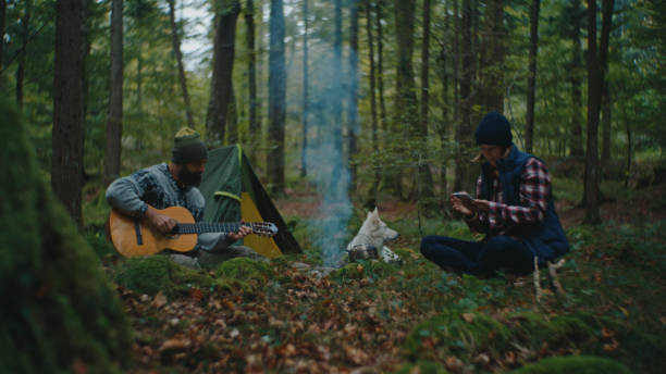 Immersed in the Forest's Tranquility,a Man's Guitar Melodies Fill the Air while his Companion Uses a Smartphone near the Campfire,Blending Modern Technology with the Timeless Charm of Outdoor Living during their Vacation.