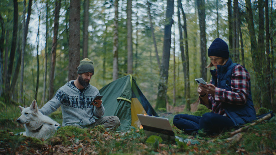 Together near the Tent in the Forest,a Man and Woman Engage with their Mobile Phones,Enjoying Digital Connectivity amidst the Tranquility of Nature during their Camping Adventure.