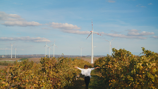 Woman Running with Pure Delight with Arms Open Wide in Gesture of Exhilaration,Making Her Way Towards the Distant Wind Turbines,Blending Seamlessly into the Serene Landscape.