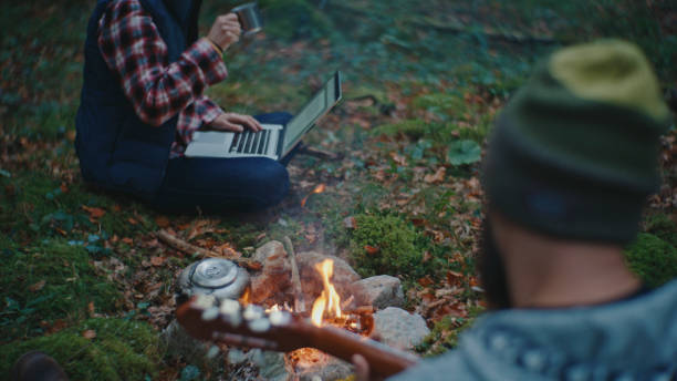 istock Man Playing Guitar while Woman Using Laptop near Campfire in Forest 2025405993