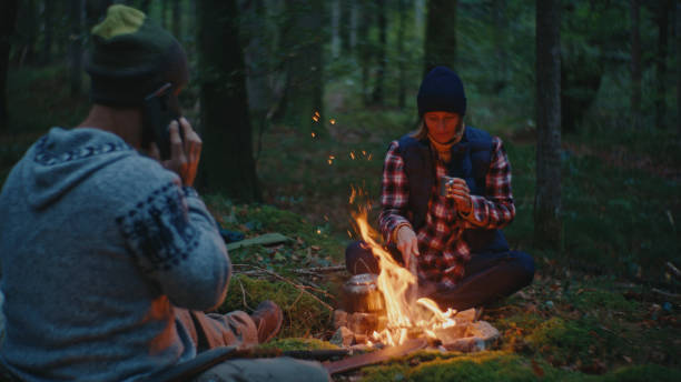 istock Man Talking on Phone while Woman Setting Up Fire at Campfire in Forest 2025407092