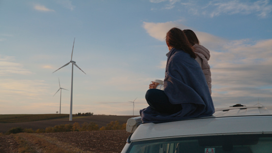 Female Friends Sitting on Car Roof with the Gentle Hum of Wind Turbines in the Vast Landscape,Surrounded by the Rhythmic Dance of the Turbines Against the Sky's Canvas.