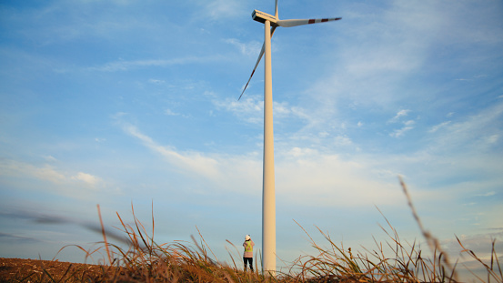 Female Engineer Standing Beside Towering Wind Turbine on Vast Agricultural Field,with Sky in Background. She Examining the Structure,Perhaps Ensuring Its Proper Functioning for Harnessing Wind Energy.