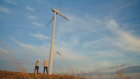 Two Women Engineers,Dressed in Work Attire,Stand Beside a Towering Wind Turbine on a Vast Agricultural Field. They Appear Focused,Analyzing the Structure Against the Backdrop of the Clear Sky,Suggesting Their Involvement in Renewable Energy Technology and Agricultural Development.