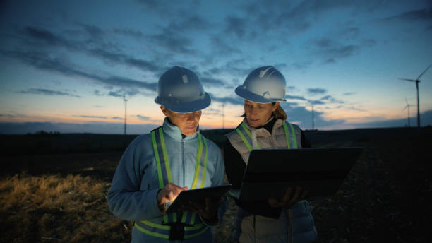 istock Female Engineers in Work Clothes Using Laptop and Tablet Talking about Wind Turbines on a Farm at Sunset 2025408229