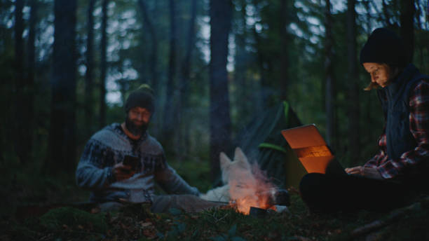 istock Woman Working on Laptop while Man Using Mobile Phone near Campfire in Forest 2025408769