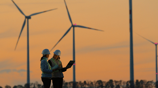 Women Engineers in Work Attire Collaborating and Utilizing Digital Tablet and Laptop Discussing the Functionality of Wind Turbines on Farm In the Serene Backdrop of Sunset.