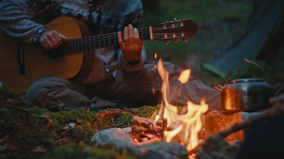Engaging in Leisurely Pursuits,a Man Spends Quality Time Playing Guitar near the Campfire in the Forest during their Vacation,Adding a Touch of Musical Charm to the Serene Woodland Setting.