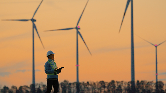 Female Engineer With the Aid of Digital Tablet Carefully Examining Wind Turbines on Farm as the Sun Sets in the Background,Ensuring Their Efficiency and Functionality.