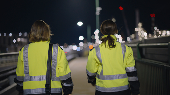 In the Gleaming City Night,Female Engineers in Reflective Clothing,Walking Across Brightly Illuminated Bridge,Showcasing Their Presence and Dedication to Their Profession.