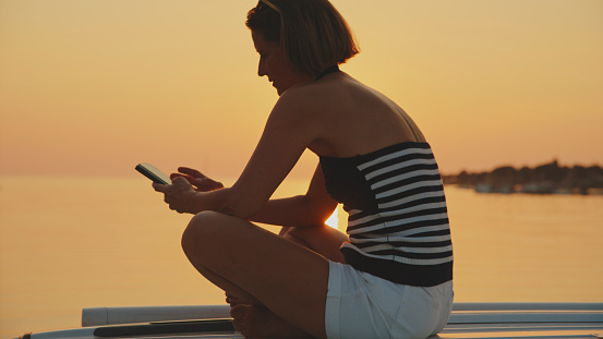 Woman Sit on Top of Van is using her Mobile Phone. she Finds Comfort and Connection,Losing Herself in the Beauty of the Peaceful Evening by the Shore. she is on Vacation.