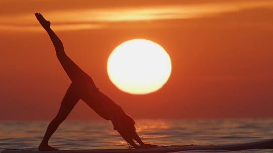 In the Ethereal Glow of the Golden Hour,a Fit Woman Gracefully Finds her Balance. Female is Practicing Yoga Atop a Paddleboard in the Tranquil Sea. Serene Pose Reflects her Inner Strength and Connection to the Rhythmic Flow of Nature.