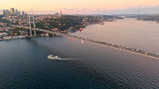 In the Enchanting Glow of Sunset at Istanbul,Turkey,an Aerial Drone View Captures a Ferry Boat Gracefully Traversing the Marmara Sea Beneath the Majestic Bosphorus Bridge,with the Cityscape as a Backdrop. The Scene Radiates with Warm Hues,Casting a Mesmerizing Ambiance Over the Urban Landscape
