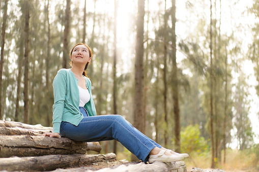 Happy beautiful Asian middle adult age woman enjoy in the nature environment in forest  with beautiful sunlight behind the trees. Leisure activity with a nature concept. Delightful Asian woman relaxing alone in the nature environment portrait.