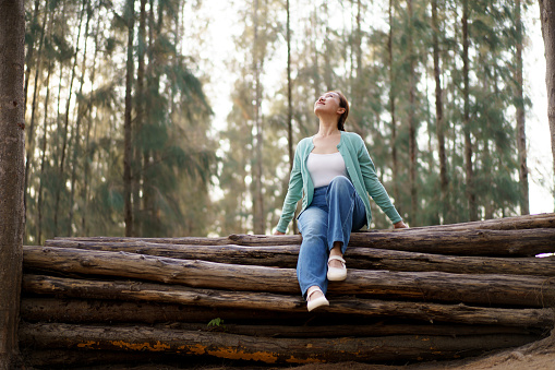 Happy beautiful Asian middle adult age woman enjoy in the nature environment in forest  with beautiful sunlight behind the trees. Leisure activity with a nature concept. Delightful Asian woman relaxing alone in the nature environment portrait.