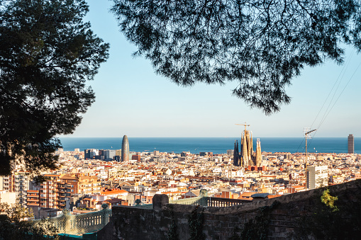 Barcelona, Spain - September 22, 2021: View through the trees from Park Guell over the city. Aerial View Cityscape with cloudy sky. Surrounded by trees with green leaves