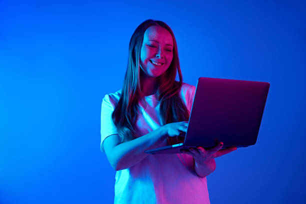 istock Attractive young woman holds online meeting using laptop against gradient blue studio background in neon light. Concept of freelance 2025467741