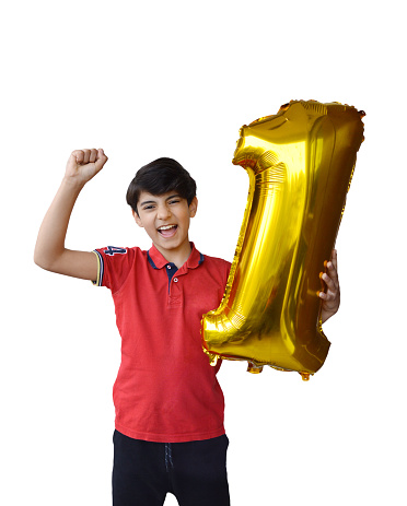 Cutout of 11 year old happy joyful energetic enthusiastic excited full of vigour child in red t shirt holding a golden shiny 3 D number1 or one foil balloon for success, win, 1st happy birthday celebrations isolated over vertical white background