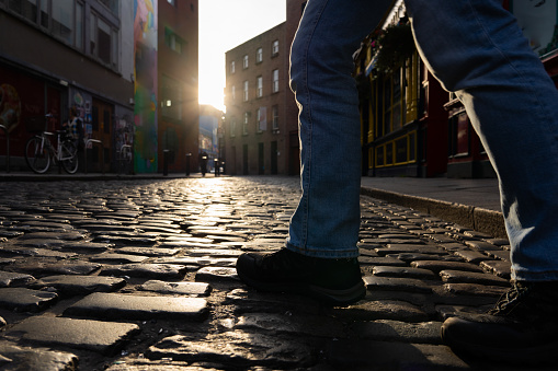 Close-up of pedestrian legs crossing a street with old paving stone on a beautiful, central street in Dublin with sun setting in the background of the buildings