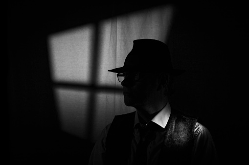 Film Noir style detective wearing a Fedora hat, tie and waistcoat in a dark, seedy office with a stylized window behind. Face partially obscured by the hat brim.