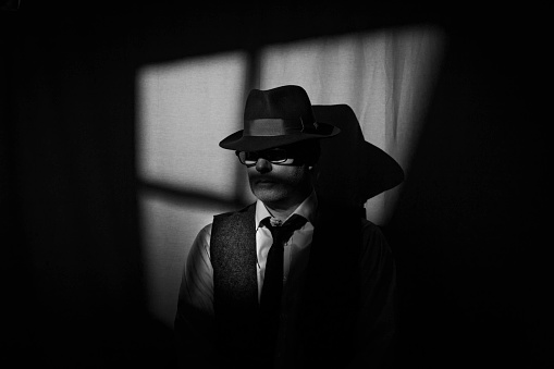 Film Noir style detective wearing a Fedora hat, tie and waistcoat in a dark, seedy office with a stylized window behind. Face partially obscured by the hat brim.
