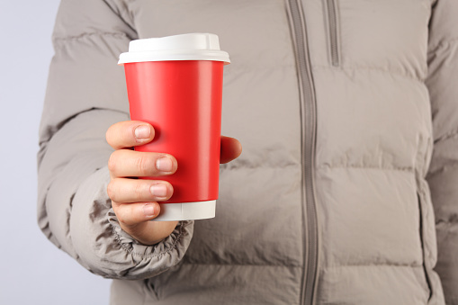 Man in a down jacket holds paper red coffee cup on a gray background
