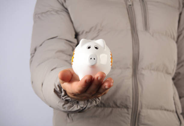 Man in down jacket holds white ceramic piggy bank on gray background.